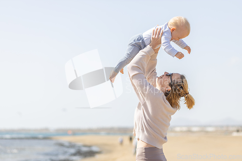 Image of Mother enjoying summer vacations holding, playing and lifting his infant baby boy son high in the air on sandy beach on Lanzarote island, Spain. Family travel and vacations concept.