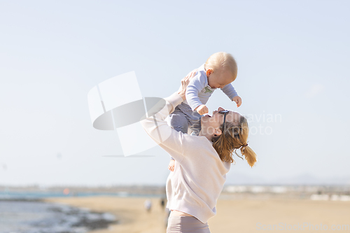 Image of Mother enjoying summer vacations holding, playing and lifting his infant baby boy son high in the air on sandy beach on Lanzarote island, Spain. Family travel and vacations concept.