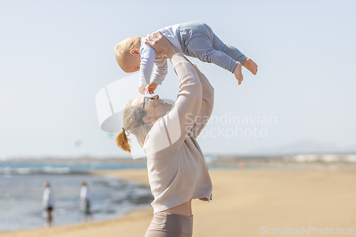 Image of Mother enjoying summer vacations holding, playing and lifting his infant baby boy son high in the air on sandy beach on Lanzarote island, Spain. Family travel and vacations concept.