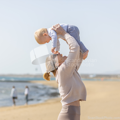 Image of Mother enjoying summer vacations holding, playing and lifting his infant baby boy son high in the air on sandy beach on Lanzarote island, Spain. Family travel and vacations concept.