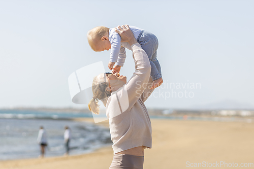 Image of Mother enjoying summer vacations holding, playing and lifting his infant baby boy son high in the air on sandy beach on Lanzarote island, Spain. Family travel and vacations concept.