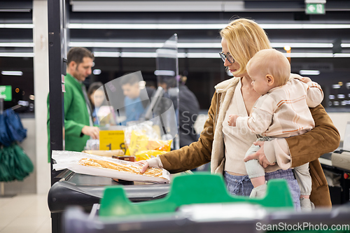 Image of Mother shopping with her infant baby boy, holding the child while stacking products at the cash register in supermarket grocery store.