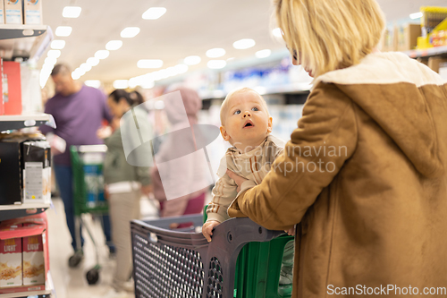 Image of Casualy dressed mother choosing products in department of supermarket grocery store with her infant baby boy child in shopping cart.