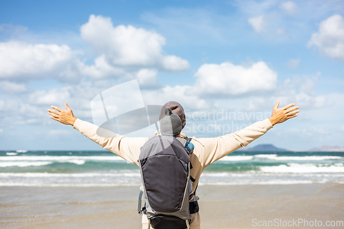 Image of Young father rising hands to the sky while enjoying pure nature carrying his infant baby boy son in backpack on windy sandy beach of Famara, Lanzarote island, Spain. Family travel concept.