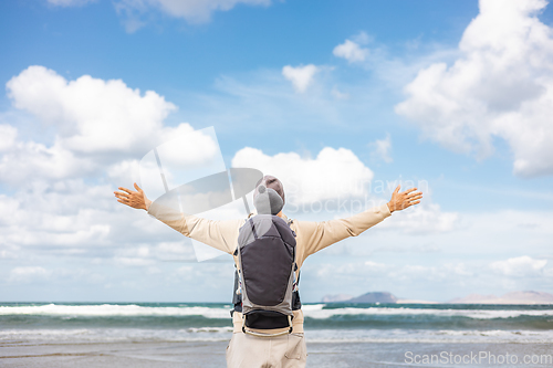 Image of Young father rising hands to the sky while enjoying pure nature carrying his infant baby boy son in backpack on windy sandy beach of Famara, Lanzarote island, Spain. Family travel concept.