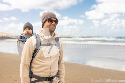 Image of Young father carrying his infant baby boy son in backpack on windy sandy beach. Family travel and winter vacation concept.