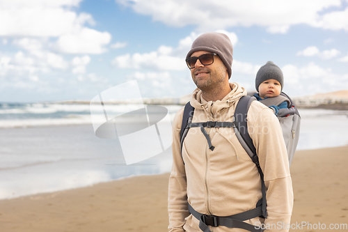 Image of Young father carrying his infant baby boy son in backpack on windy sandy beach. Family travel and winter vacation concept.