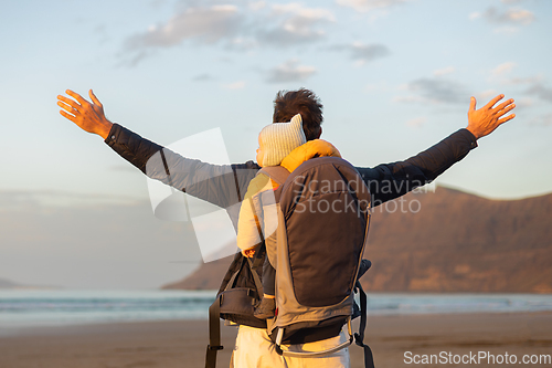 Image of Young father rising hands to the sky while enjoying pure nature carrying his infant baby boy son in backpack on windy sandy beach of Famara, Lanzarote island, Spain at sunset. Family travel concept.