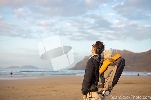 Image of Young father rising hands to the sky while enjoying pure nature carrying his infant baby boy son in backpack on windy sandy beach of Famara, Lanzarote island, Spain at sunset. Family travel concept.