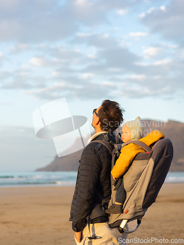 Image of Young father rising hands to the sky while enjoying pure nature carrying his infant baby boy son in backpack on windy sandy beach of Famara, Lanzarote island, Spain at sunset. Family travel concept.