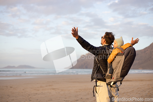 Image of Young father rising hands to the sky while enjoying pure nature carrying his infant baby boy son in backpack on windy sandy beach of Famara, Lanzarote island, Spain at sunset. Family travel concept.