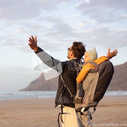 Image of Young father rising hands to the sky while enjoying pure nature carrying his infant baby boy son in backpack on windy sandy beach of Famara, Lanzarote island, Spain at sunset. Family travel concept.