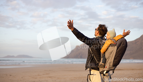 Image of Young father rising hands to the sky while enjoying pure nature carrying his infant baby boy son in backpack on windy sandy beach of Famara, Lanzarote island, Spain at sunset. Family travel concept.