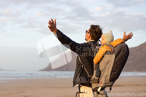 Image of Young father rising hands to the sky while enjoying pure nature carrying his infant baby boy son in backpack on windy sandy beach of Famara, Lanzarote island, Spain at sunset. Family travel concept.