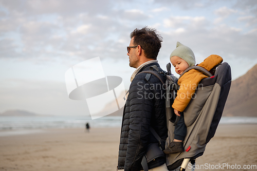 Image of Father enjoying pure nature carrying his infant baby boy son in backpack on windy sandy beach of Famara, Lanzarote island, Spain at sunset. Family travel concept.