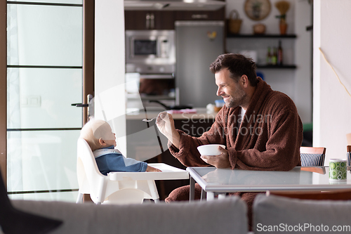 Image of Father wearing bathrope spoon feeding hir infant baby boy child sitting in high chair at the dining table in kitchen at home in the morning.