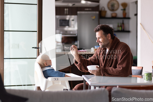 Image of Father wearing bathrope spoon feeding hir infant baby boy child sitting in high chair at the dining table in kitchen at home in the morning.