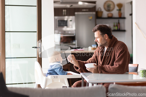 Image of Father wearing bathrope spoon feeding hir infant baby boy child sitting in high chair at the dining table in kitchen at home in the morning.