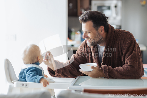 Image of Father wearing bathrope spoon feeding hir infant baby boy child sitting in high chair at the dining table in kitchen at home in the morning.