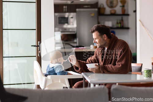 Image of Father wearing bathrope spoon feeding hir infant baby boy child sitting in high chair at the dining table in kitchen at home in the morning.