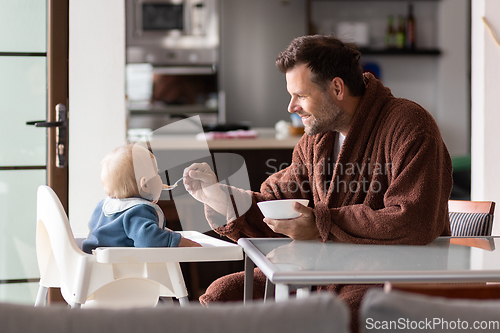 Image of Father wearing bathrope spoon feeding hir infant baby boy child sitting in high chair at the dining table in kitchen at home in the morning.