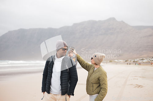 Image of Young father carrying his infant baby boy son in backpack on windy sandy beach. Family travel and winter vacation concept.