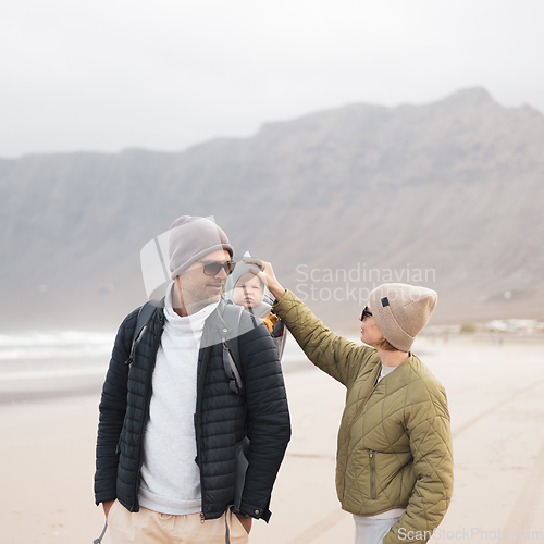 Image of Young father carrying his infant baby boy son in backpack on windy sandy beach. Family travel and winter vacation concept.