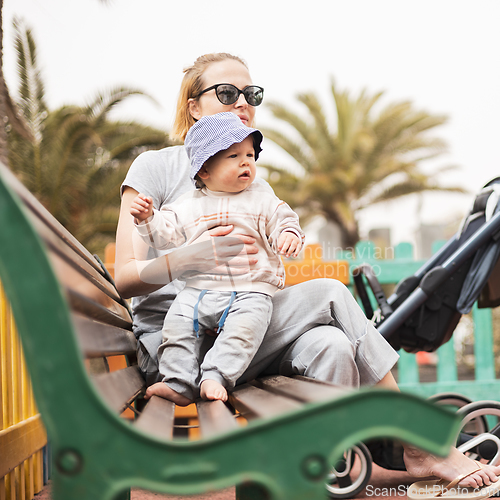 Image of Young mother with her cute infant baby boy child on bench on urban children's playground on warm summer day.