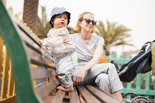 Image of Young mother with her cute infant baby boy child on bench on urban children's playground on warm summer day.