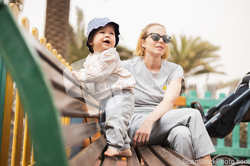 Image of Young mother with her cute infant baby boy child on bench on urban children's playground on warm summer day.