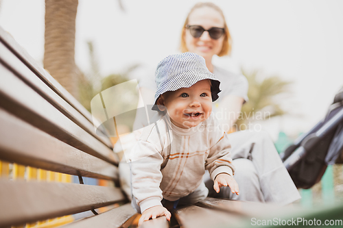 Image of Young mother with her cute infant baby boy child on bench on urban children's playground on warm summer day.