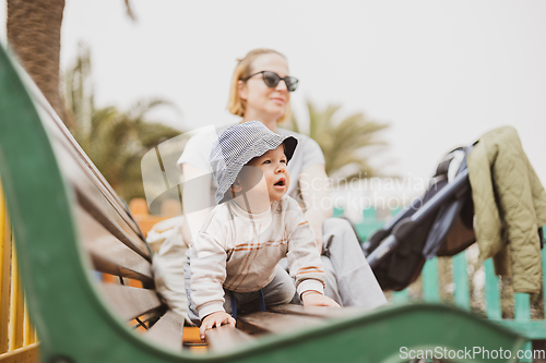 Image of Young mother with her cute infant baby boy child on bench on urban children's playground on warm summer day.