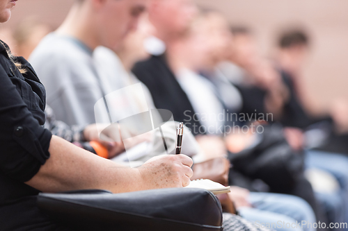 Image of Female hands holding pen and notebook, making notes at conference lecture. Event participants in conference hall.