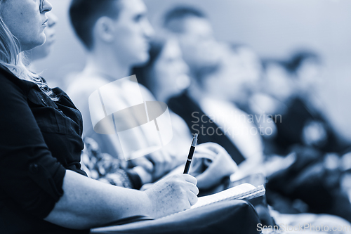Image of Female hands holding pen and notebook, making notes at conference lecture. Event participants in conference hall. Blue toned greyscale image.