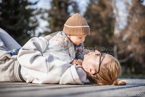 Image of Happy family. Young mother playing with her baby boy infant oudoors on sunny autumn day. Portrait of mom and little son on wooden platform by lake. Positive human emotions, feelings, joy.