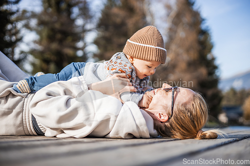 Image of Happy family. Young mother playing with her baby boy infant oudoors on sunny autumn day. Portrait of mom and little son on wooden platform by lake. Positive human emotions, feelings, joy.