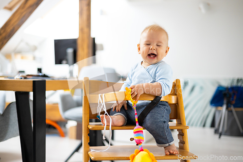 Image of Happy infant sitting and playing with his toy in traditional scandinavian designer wooden high chair in modern bright atic home. Cute baby.