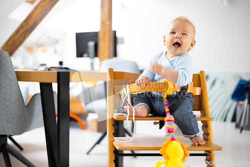 Image of Happy infant sitting and playing with his toy in traditional scandinavian designer wooden high chair in modern bright atic home. Cute baby.