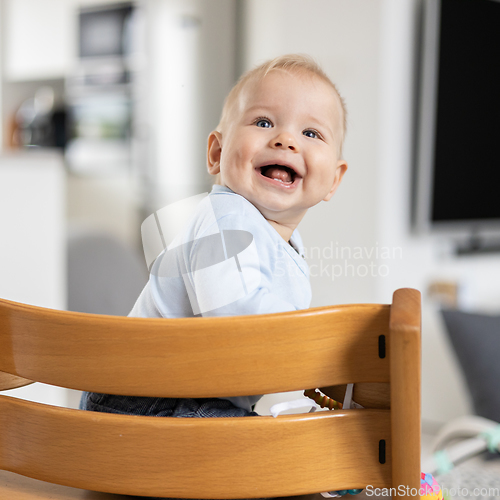 Image of Happy infant sitting in traditional scandinavian designer wooden high chair and laughing out loud in modern bright home. Cute baby smile.