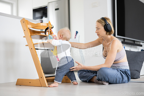 Image of Women's multitasking. Mother sitting on floor playing with her baby boy watching and suppervising his first steps while listening to podcast on wireless headphones.