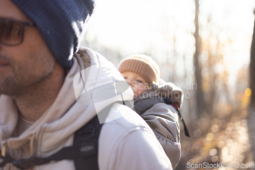 Image of Sporty father carrying his infant son wearing winter jumpsuit and cap in backpack carrier hiking in autumn forest.