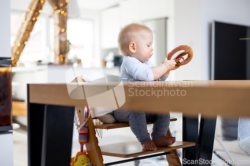 Image of Happy infant sitting at dining table and playing with his toy in traditional scandinavian designer wooden high chair in modern bright atic home. Cute baby playing with toys