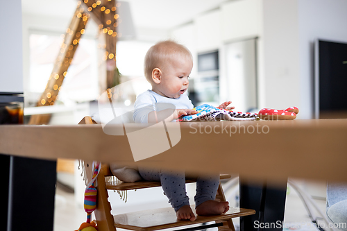 Image of Happy infant sitting at dining table and playing with his toy in traditional scandinavian designer wooden high chair in modern bright atic home. Cute baby playing with toys