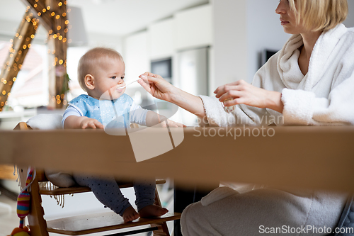 Image of Mother wearing cosy bathrope spoon feeding her baby boy child in baby high chair with fruit puree at dinning table at home. Baby solid food introduction concept