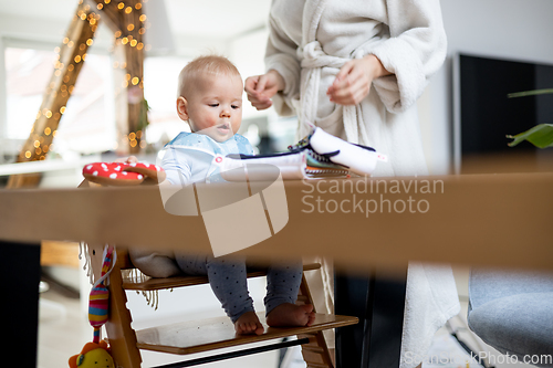 Image of Happy infant sitting at dining table and playing with his toy in traditional scandinavian designer wooden high chair in modern bright atic home superwised by his mother