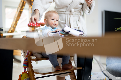Image of Happy infant sitting at dining table and playing with his toy in traditional scandinavian designer wooden high chair in modern bright atic home superwised by his mother.
