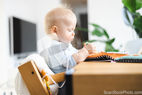 Image of Happy infant sitting at dining table and playing with his toy in traditional scandinavian designer wooden high chair in modern bright atic home. Cute baby playing with toys