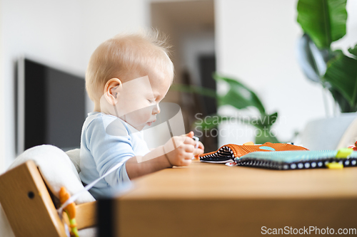 Image of Happy infant sitting at dining table and playing with his toy in traditional scandinavian designer wooden high chair in modern bright atic home. Cute baby playing with toys