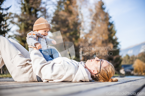 Image of Happy family. Young mother playing with her baby boy infant oudoors on sunny autumn day. Portrait of mom and little son on wooden platform by lake. Positive human emotions, feelings, joy.