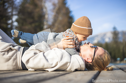 Image of Happy family. Young mother playing with her baby boy infant oudoors on sunny autumn day. Portrait of mom and little son on wooden platform by lake. Positive human emotions, feelings, joy.